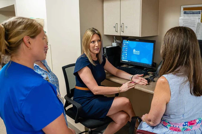 A person sitting at a desk in a medical office engages in conversation with three others. A computer screen in the background displays the MUSC Hollings Cancer Center logo.