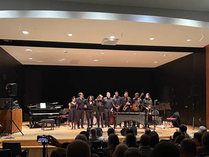 A group of musicians stands in a line on a stage, taking a bow in front of an audience. A grand piano is visible on the left, and various instruments, including a cello and violin, are held by some of the musicians. The backdrop is a minimalist, black stage setup, and audience members are seated in the foreground.