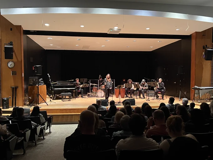 A group of musicians performs on a stage in an auditorium. There are various instruments like a piano, drums, and a double bass. The audience is seated in rows, watching attentively. The setting is well-lit, and the stage is surrounded by wooden panels.