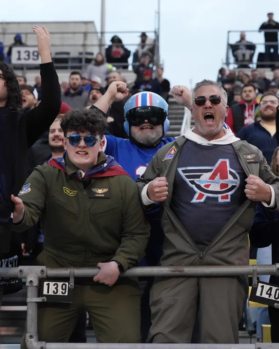 A group of enthusiastic fans dressed in aviation-themed outfits cheer loudly at a sports event, conveying excitement and support.