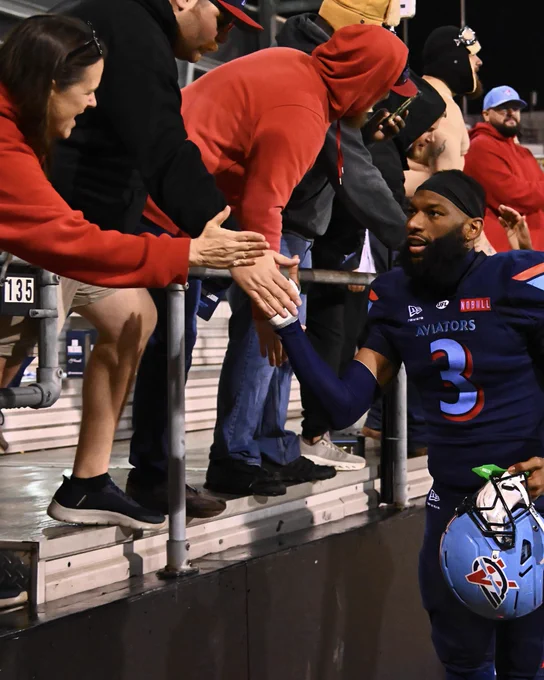 A football player from the Aviators team interacts with fans by shaking hands over a stadium barrier. The scene is warm and engaging, highlighting the connection between the player and supporters.