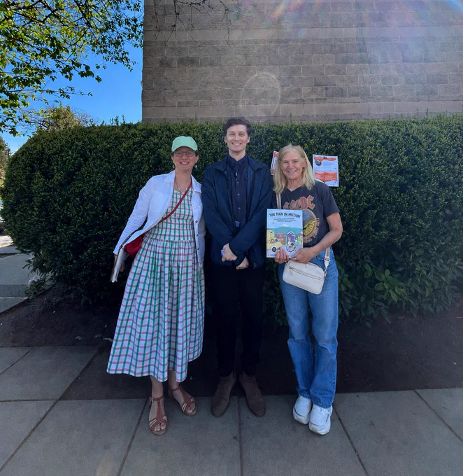 A man in a blue sweatshirt, standing between a woman in a green hat and dress, an a woman in blue jeans holding a book
