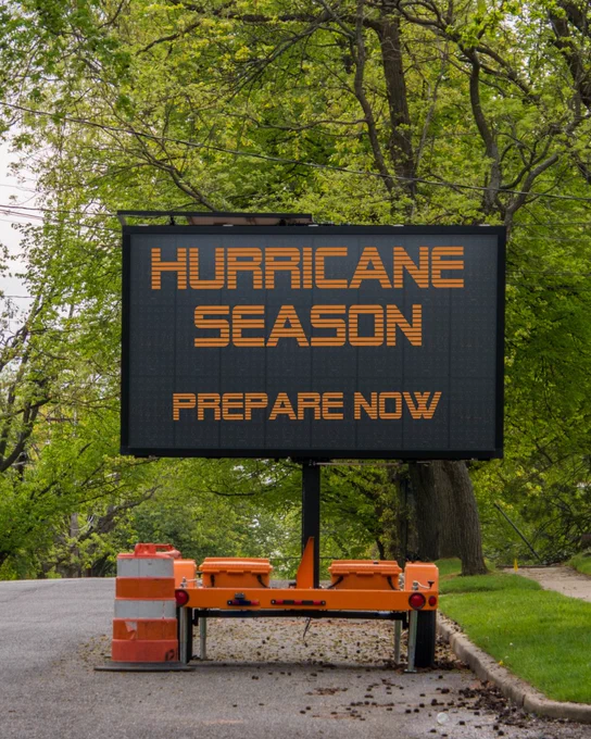 A roadside electronic sign amidst lush green trees displays the message: "Hurricane Season Prepare Now."