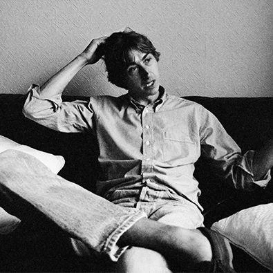 Mark Hollis of Talk Talk in a black-and-white portrait circa mid-1980s, seated casually on a sofa with one hand resting on his head, dressed in a relaxed shirt and jeans, capturing a thoughtful, understated mood reflective of the It’s My Life era.