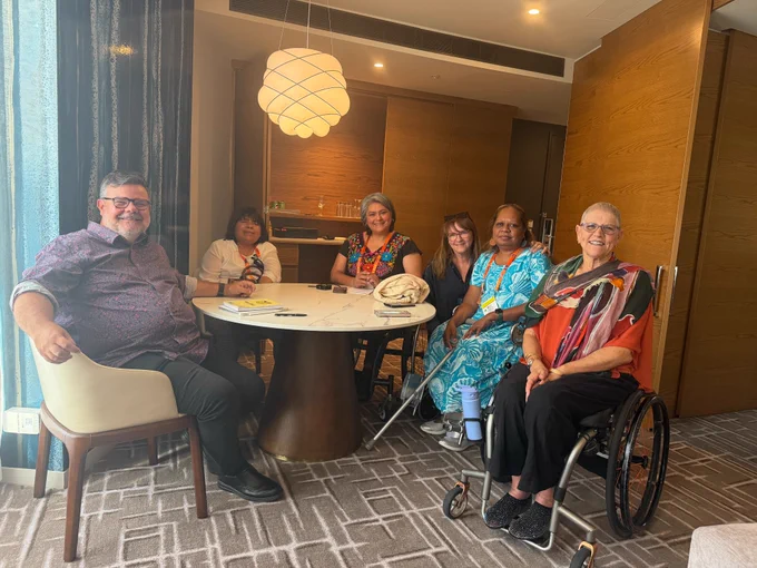 A group of six people are gathered around a round, white marble-topped table in a brightly lit room. From left to right: Damian Griffis, Rosario Galarza (Peru), Olga Montufar (Mexico), Allison Wong (FPDN), Lanieta Tuimabu (Fiji), Aunty Kay Sandler.