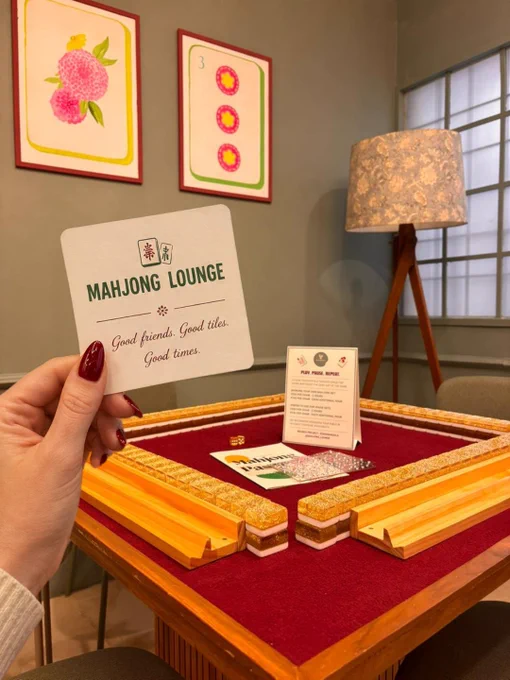 A cozy and inviting scene inside Mahjong Lounge featuring a Mahjong table with a rich red playing surface and neatly arranged wooden tile racks. In the foreground, a hand with red nail polish holds a small card that reads “Mahjong Lounge – Good friends. Good tiles. Good times.” The background includes warm ambient lighting from a floor lamp, framed artwork on the wall, and a softly lit, comfortable interior setting. Mahjong tiles and game elements are arranged on the table, creating a sense of anticipation before gameplay begins. The overall composition highlights a premium, welcoming environment designed for social gaming, where players gather to relax, connect, and enjoy Mahjong in a refined yet approachable space in Bangalore.
