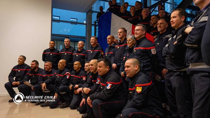 Un grand groupe de pompiers français en uniforme bleu marine avec des bandes rouges posent pour une photo de groupe à l'intérieur d'un bâtiment moderne. Ils sont rassemblés devant un drapeau tricolore bleu-blanc-rouge. Le logo 'Sécurité Civile & Gestion des Crises' figure en bas à gauche de l'image.
