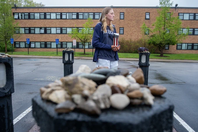 A person stands in a parking lot, holding several rolled diplomas. In the foreground, a pile of rocks is in focus. The building in the background appears to be a school or institutional facility. The setting is overcast and the ground is wet.