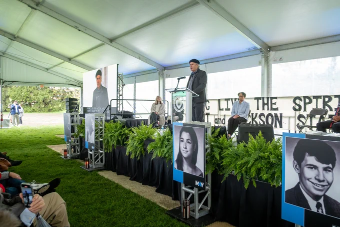 A group of people are gathered under a large tent canopy for an event. A speaker stands at a podium adorned with the word "LONG" next to images of young individuals. Fern plants decorate the stage area. Attendees are seated and focused on the presentation.