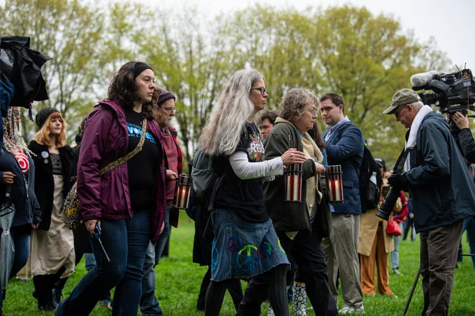 A group of people holding lanterns are gathered in at Kent State, with trees in the background. A person with a camera and microphone stands nearby, capturing the event. The mood seems reflective and purposeful.