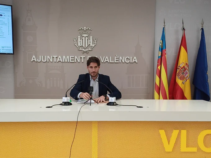 El portavoz municipal, Juan Carlos Caballero, explica los acuerdos de la Junta de Gobierno Local en la sala de prensa de Ayuntamiento de València. Está sentado, con con micrófonos delante y banderas de la Comunidad Valenciana, España y UE detrás.