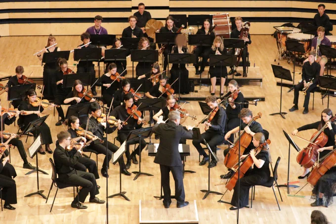 Conductor leading a full orchestra with string, woodwind, and percussion sections on a wooden stage in a concert hall.