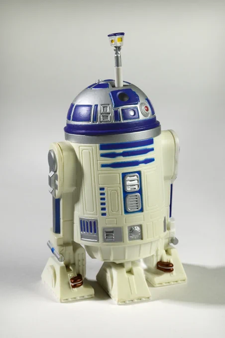 A toy replica of R2-D2 from Star Wars, featuring a metallic dome, blue and white body with various panels and a small antenna on top, standing on a plain surface.