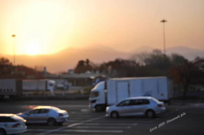 ALT :  ⬇️  

A slightly blurred, out-of-focus photograph of an SA (Service Area) parking lot at sunrise. The sunlight from the daybreak is a bright, hazy spot over rolling hills in the background, while the shapes of large trucks and passenger cars are softened by the shallow depth of field.

Location: Iwatesan Service Area (Northbound)
Hirakasa, Hachimantai, Iwate-pref. 

©2026 MRP. Yamamoto | photographié