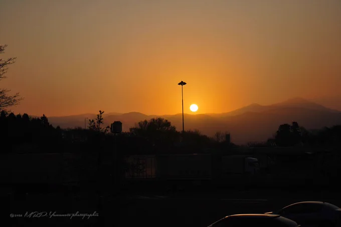 ALT :  ⬇️

A landscape photograph of a bright sunrise positioned behind a silhouette of mountains during daybreak early morning hours. In the dark foreground, the silhouettes of parked trucks, cars, and a tall light pole are visible under an orange and yellow sky.

Location: Iwatesan Service Area (Northbound)
Hirakasa, Hachimantai, Iwate-pref. 

©2026 MRP. Yamamoto | photographié