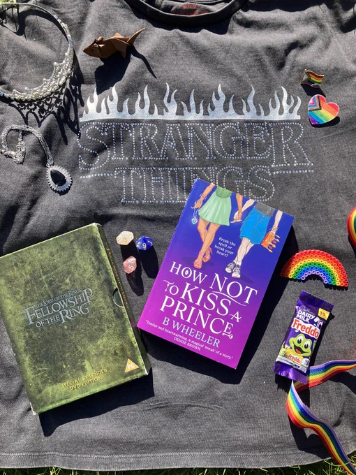 Background of grey Stranger Things t-shirt. Paperback of How NOT To Kiss A Prince by B Wheeler surrounded by assorted items. Clockwise from top left: a tiara, origami rat, 2 pride pines, a beaded rainbow, a Cadbury’s Freddo, rainbow ribbon,The Lord of the Rings DVD boxset, a silver necklace with a blue pendant.