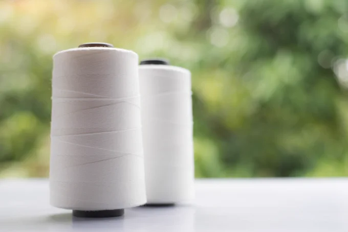 Two white spools of thread sit upright on a light surface, one in sharp focus in the foreground and one slightly blurred behind it, against a soft green, out‑of‑focus background suggesting foliage or nature.