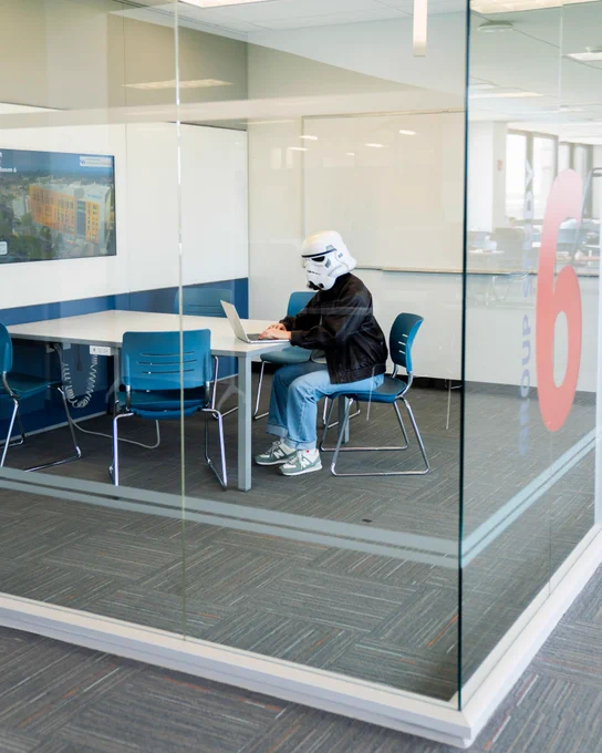 Student types on their laptop in a group study room while wearing a Storm Trooper helmet