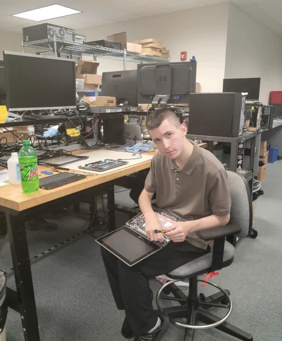 A person is seated on a stool in a computer repair workshop, holding and working on an open laptop. The desk is cluttered with electronic equipment and a bottle of Mountain Dew. Computer monitors and other devices are visible in the background.
