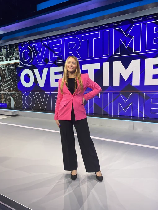 Photo of a girl posing standing with her hands on her hips, wearing black pants and shoes and a pink blazer. She has blonde hair and is smiling. In the background there is a wall in blue and white letters that says overtime.