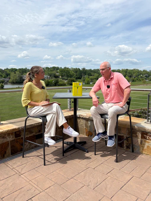 Photo of a woman interviewing a man outside and they are both sitting at a table. The woman is wearing a yellow sweater and white pants with white sneakers and blonde hair. The man is wearing a pink shirt, khaki pants, and blue sneakers. There is a book on the table that says All Cary with a yellow cover and a golf club and golf ball on it. There are blue skies behind them and clouds with trees and a lake in the background.