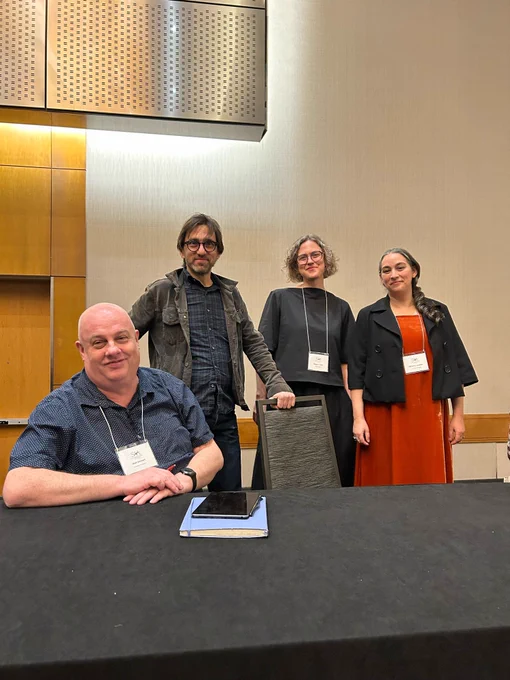 One panelist sits at a table while three other panelists stand behind him. End ID.