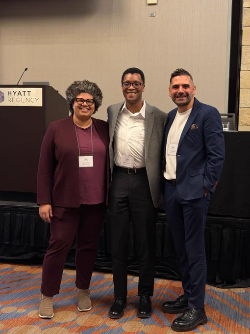 Image of three panelists smiling in front of a stage and podium. End ID.