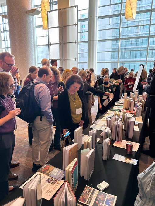 Image of a crowd of conference-goers examining a table covered in newly released hardcover books. End ID.
