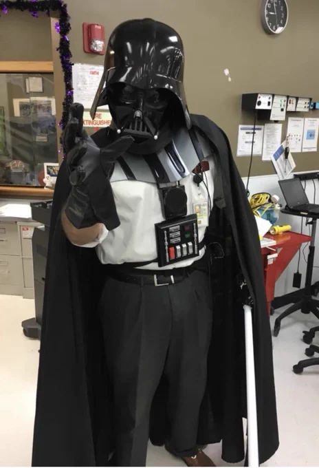 Photo of me wearing my Darth Vader helmet and cloak over my clinic business attire, with a lightsaber in one gloved hand and the other one making a “Force choke” gesture towards the camera.  I’m standing in my office.