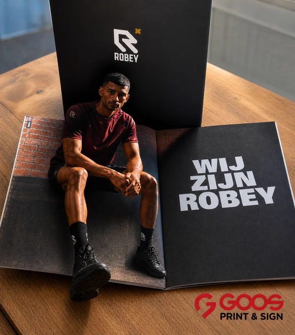 Een open luxe magazine van sportkledingmerk Robey ligt op een houten tafel. Op de spread zit een sportieve man in bordeauxrode sportkleding en zwarte sneakers. Op de rechterpagina staat in grote witte letters “WIJ ZIJN ROBEY”. Onderaan de afbeelding staat het logo van GOOS Print & Sign.