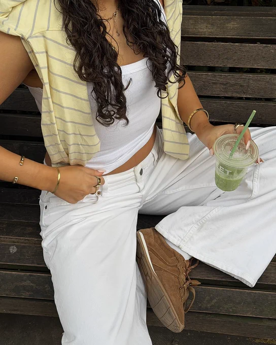 An image of a person wearing a pair of dark brown Steve Madden Artiste Ballet trainers with a white outfit whilst sitting on a wooden bench holding a green drink.