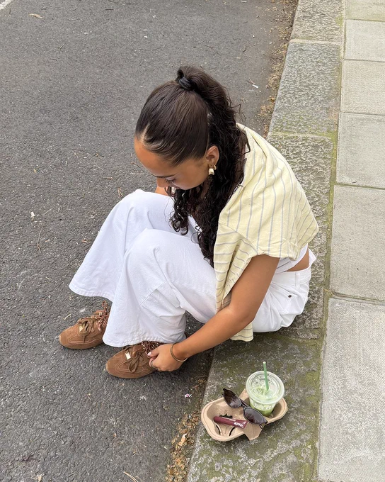 An image of a person wearing a pair of dark brown Steve Madden Artiste Ballet trainers with a white outfit whilst sitting on a roadside next to a green drink.
