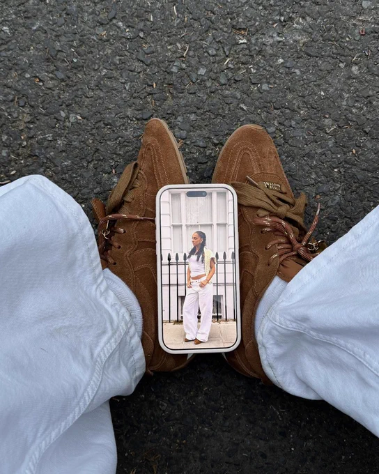 An image of a person's lower legs wearing a pair of dark brown Steve Madden Artiste Ballet trainers whilst sitting on a roadside with a mobile phone resting on their feet. 