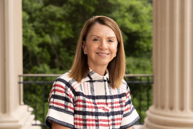 Portrait of a woman with straight light brown shoulder-length hair outside on a collegiate building's porch with columns. 