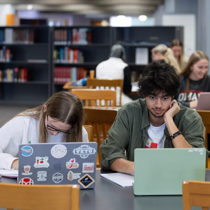 Two students studying. There is a woman with long hair looking at a notebook, and a man with brown hair looking at a laptop screen. In the background there are book shelves and other students studying.