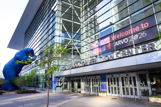 An exterior view of the Colorado Convention Center with a large blue bear sculpture peering inside. A digital sign reads "Welcome to ARVO 2026." Modern glass facade and nearby trees are visible.
