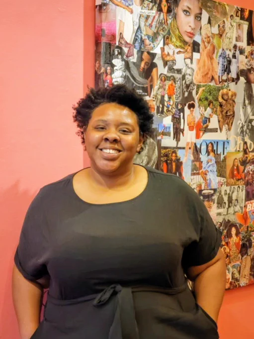 Danielle Demetria East tilts her head slightly and smiles at the camera. She wears a black empire waist dress and has her hands in her pockets. She stands in front of a large photo collage on a coral-colored wall. Photo: Lee-Vi Salinas