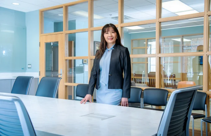 A woman stands confidently at a large conference table in a modern office space. She wears a black blazer over a light blue shirt and has long dark hair. The room features glass walls and is organized with chairs around the table, conveying a professional atmosphere.