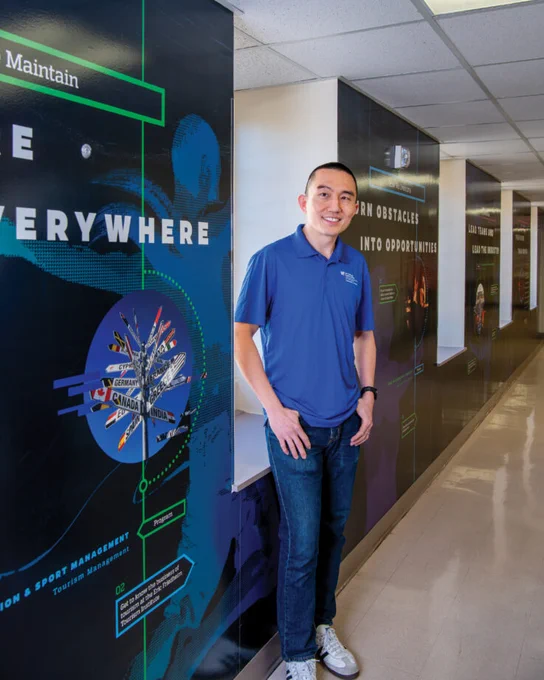 A man stands in a corridor with educational posters on the walls. He is wearing a blue polo shirt and jeans, smiling at the camera. The hallway features graphics related to various fields of study or career opportunities.