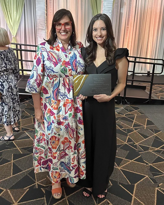Two individuals are smiling for a photo indoors. One is wearing a colorful patterned dress and glasses, and the other is in a black outfit, holding a black folder. The background features draped curtains and geometric-patterned carpet.