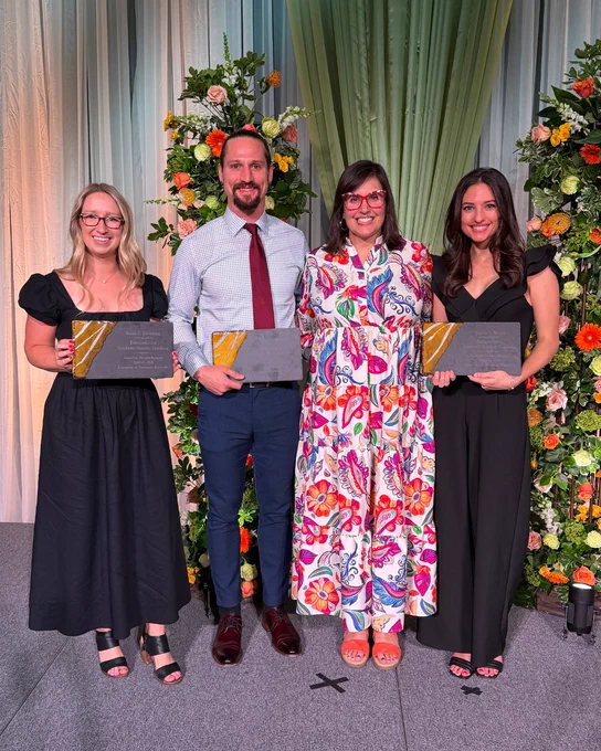 Four individuals stand on a stage holding plaques. They are dressed formally, with floral arrangements in the background.