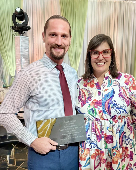 Two people smiling and standing together, with one holding an award plaque for "Best Shelton Experimentalist Graduate Student Teacher.