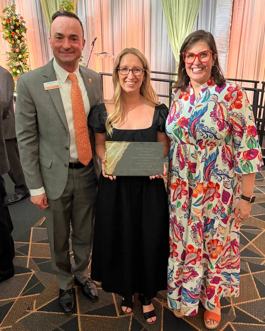 Three individuals are standing indoors, dressed in formal attire. The person in the center holds a plaque. Two are wearing name tags, and one wears a vibrant, patterned dress. The background features draped fabric and stage equipment.