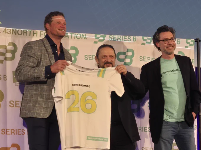 Three people stand on a stage in front of a branded backdrop reading \u201CBuffalo Startup Week\u201D and \u201CSeries B,\u201D holding up a white jersey with the number \u201C26.\u201D The stage is lit with blue lighting, and the backdrop features repeated \u201CSeries B\u201D and \u201C43North Foundation\u201D logos.