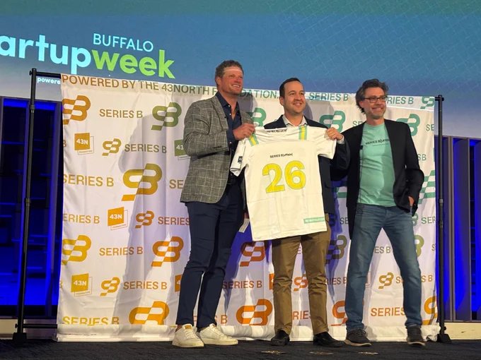Close-up of three people on stage holding a white jersey labeled \u201CSeries B Buffalo\u201D with the number \u201C26,\u201D standing in front of a backdrop covered with \u201CSeries B\u201D logos and event branding.