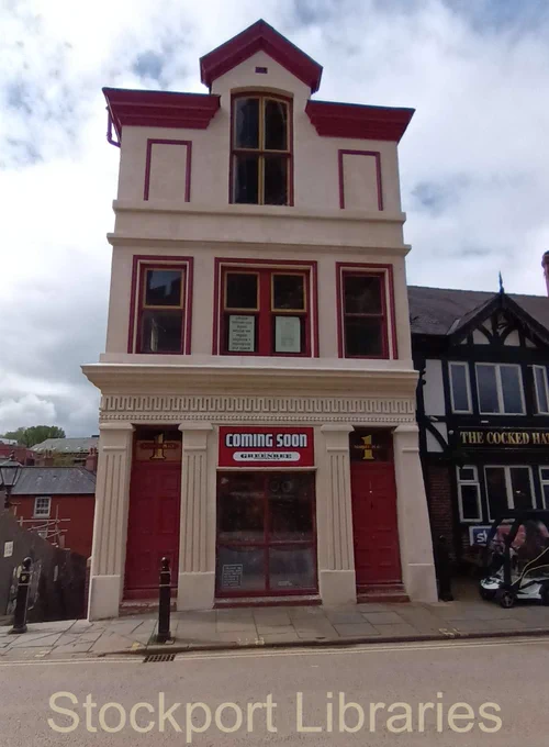 Three storey building with a central window on the second floor with a gabled roof. To the ground floor, two doors one to the left and one to the right, with a central window. Next door to the right is a black and white pub building.