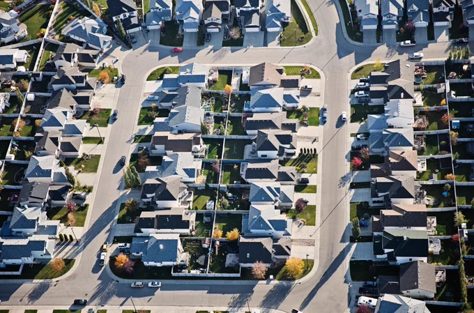 Aerial view of a suburban neighbourhood with rows of houses, driveways, and intersecting streets under clear daylight.
