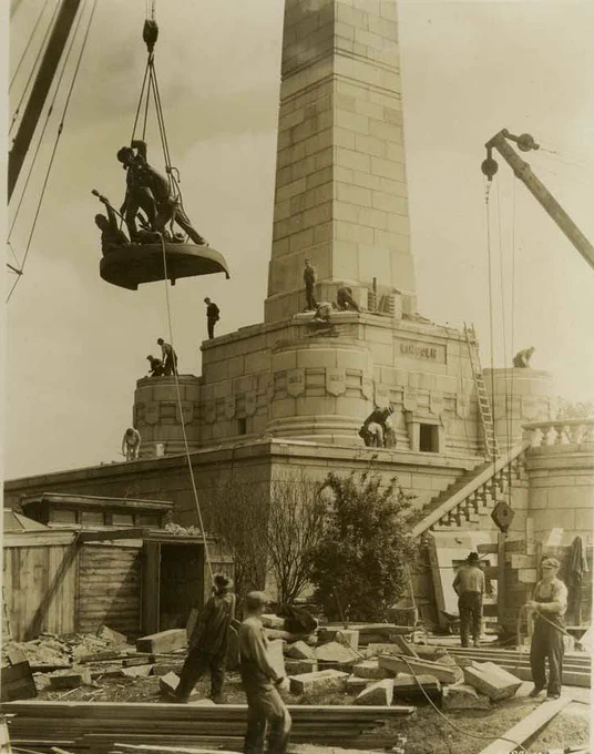 A photo of workers using cranes to install a large statue atop Lincoln's tomb under construction with scaffolding and building materials scattered below.