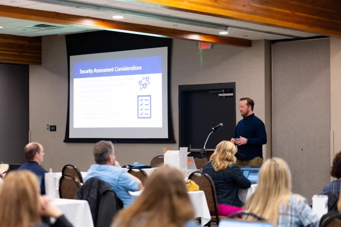 Man presenting security assessment considerations to an audience in a conference room with a projector screen.