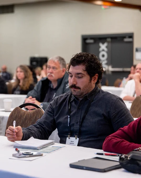 Man attentively taking notes during a conference session in a crowded meeting room.